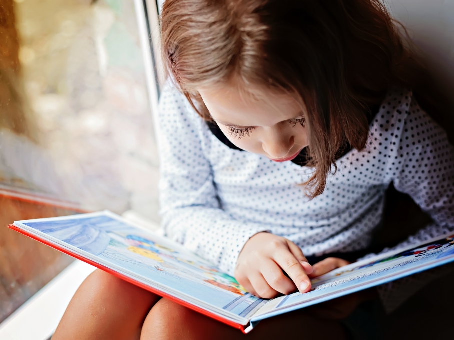 Young girl reading a book while sat on a window sill seat