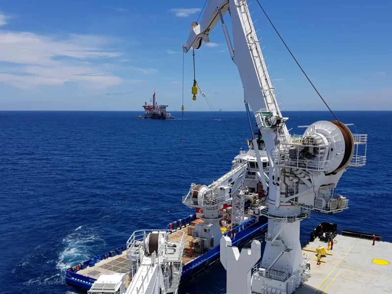 Cargo being shifted between ships at busy oilfield with use of large cranes and mechanical sheaves.